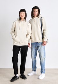 Beige hoodies med framfickor, svarta byxor och denimjeans med broderade lappar, kompletterade av svarta och vita sneakers.