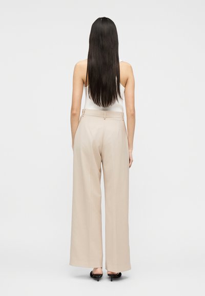 Γυναίκα που στέκεται με την πλάτη, φοράει μπεζ παντελόνι με φαρδιά μπατζάκια, λευκό αμάνικο τοπ, μαύρα ψηλοτάκουνα παπούτσια, με μακριά ίσια σκούρα μαλλιά.