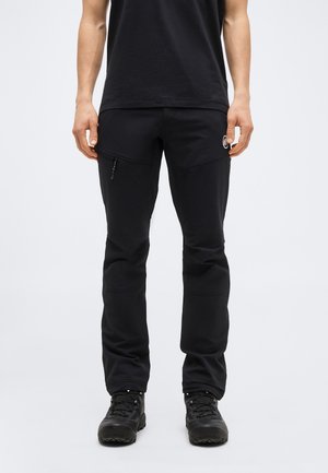 Άνδρας φορώντας μαύρο υπαίθριο παντελόνι με τσεπάκι με φερμουάρ και μαύρα παπούτσια πεζοπορίας, που στέκεται μπροστά σε απλό φόντο.