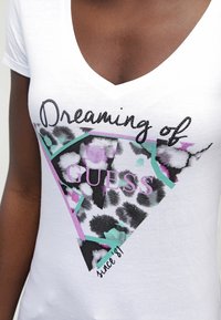 Detalle de una camiseta blanca de cuello en V con un triángulo de estampado de leopardo en negro, rosa y turquesa y el texto "Soñando con GUESS desde '81" en el frente.