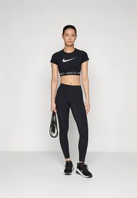 Mustad Nike Pro lühike t-särk valge logoga, koos mustade kõrge vöökohaga retuuside ja mustade spordijalatsitega, hoides hüppenööri.