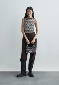 Svarta knähöga stövlar, mönstrad ärmlös topp, lösa bruna byxor med zickzackmönster och en liten flermönstrad handväska.