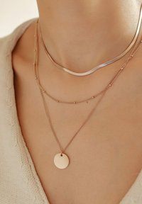 Trzy warstwowe naszyjniki: cienki srebrny łańcuszek, delikatny łańcuszek w kolorze różowego złota z małymi koralikami oraz naszyjnik z różowego złota z okrągłym medalionem.