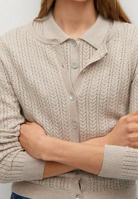 Beige neulottu neuletakki, jossa on teksturoitu, kaapelikuosinen kuvio, kauluksellinen pääntie ja nappikiinnitys. Pitkät hihat, joissa resorit hihansuissa.