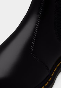 Bota de cano curto em pele preta com textura suave, costura amarela e painel de malha. O design destaca uma silhueta elegante e minimalista.