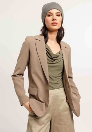 Frau trägt eine graue Mütze, ein beige Blazer, ein olivgrünes, drapiertes Oberteil und lockere beige Hosen, die Hände in den Taschen, vor einem schlichten Hintergrund.