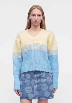 Kvinde med langt blondt hår iført en pastelgul, grå og blå stribet trøje og et blåt denimskørt med store polkaprikker.
