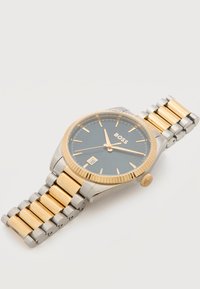 Ανδρικό ρολόι χειρός με χρυσό και ασημένιο μεταλλικό λουράκι, μαύρο καντράν, χρυσούς δείκτες και δείκτες ώρας, ένδειξη ημερομηνίας στις 6 και το λογότυπο "BOSS" στο καντράν.