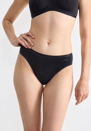 Vrouw in zwarte hooggesneden slips en een bijpassende zwarte bh, staande met één hand op de heup tegen een vlakke lichte achtergrond.