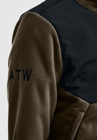Kurtka polarowa w oliwkowej zieleni z gładką czarną panelfazą na ramionach. Posiada wyszywane logo "ATW" w czerni na rękawie.
