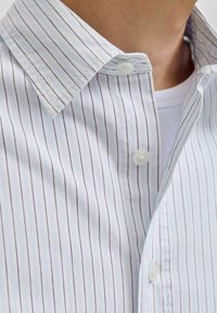 Hvid skjorte med fine lodrette sorte og lyseblå striber over en hvid t-shirt med rund hals, delvist åben i kraven.