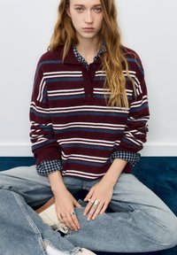 Bluza tricotată cu dungi burgund cu guler cu nasturi, purtată peste o cămașă în carouri. Blugi bleu deschis și adidași albi completează ținuta.