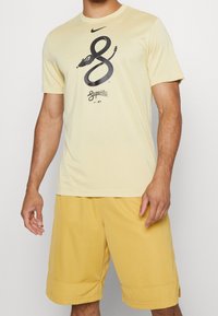 Camiseta de Nike de color amarillo con mangas cortas, que presenta un gráfico de una serpiente negra y texto. Combinada con pantalones cortos texturizados amarillos, tanto informales como atléticos.