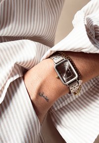 Montre rectangulaire en argent avec un cadran gris, un bracelet en métal à maillons et un pendentif accentué d'or. Une chemise rayée et un tatouage au poignet sont visibles.