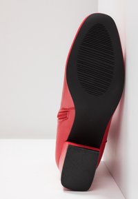 Chaussure en cuir rouge avec semelle en caoutchouc noire texturée et talon carré, positionnée debout contre un mur blanc d'angle.