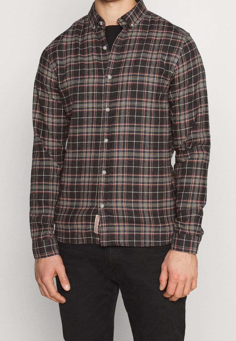 Homme portant une chemise à carreaux noire, rouge et blanche à manches longues boutonnée, avec un pantalon noir, debout sur un fond clair uni.