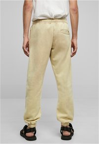Beige joggebukser med snor i livet, elastiske mansjetter og en baklomme. Brukt med svarte sandaler. Glatt tekstur, avslappet passform.