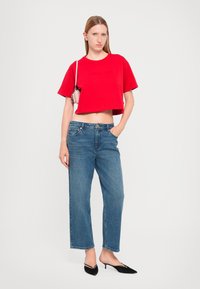 Tricou roșu scurt, blugi albaștri lejeri, tocuri negre cu prindere pe spate. Modelul poartă o geantă mică tip crossbody; stil casual cu detalii hardware minime.