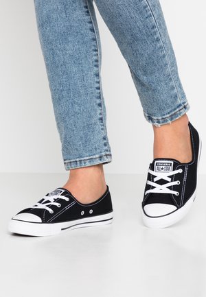 Person iført sorte lave Converse All Star sneakers med hvide snørebånd og såler, kombineret med opkørte lyseblå jeans.