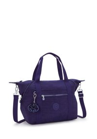 Fioletowa torba wykonana z materiału, z dwiema górnymi uchwytami, odczepianym paskiem na ramię, przednią kieszenią na zamek, okrągłym logo oraz puszystym breloczkiem.
