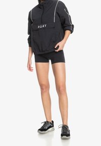 Svart vindjacka med hög krage, grå detaljer och framficka, matchad med svarta shorts och svarta sneakers med silverfärgade snörningar.