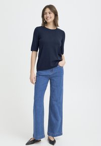 Tengerészkék rövid ujjú kötött felső, világoskék bő szárú farmerrel és fekete ráhúzható cipővel, egyszínű fehér háttér előtt állva.