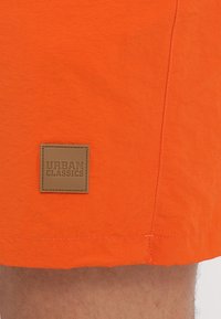 Svijetloplave kupaćice od nabrane tkanine s kvadratnom smeđom kožnom etiketom na kojoj je u izvedbi reljefa ispisano "URBAN CLASSICS".