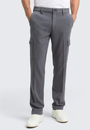 Uomo che indossa pantaloni cargo grigi a gamba dritta con tasche laterali e scarpe da ginnastica bianche, in piedi con le mani in tasca.