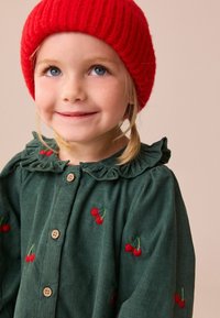 Groene corduroy blouse met een geraffineerde kraag, versierd met rode kersborduursels en houten knopen. Een rode gebreide muts zorgt voor contrast.