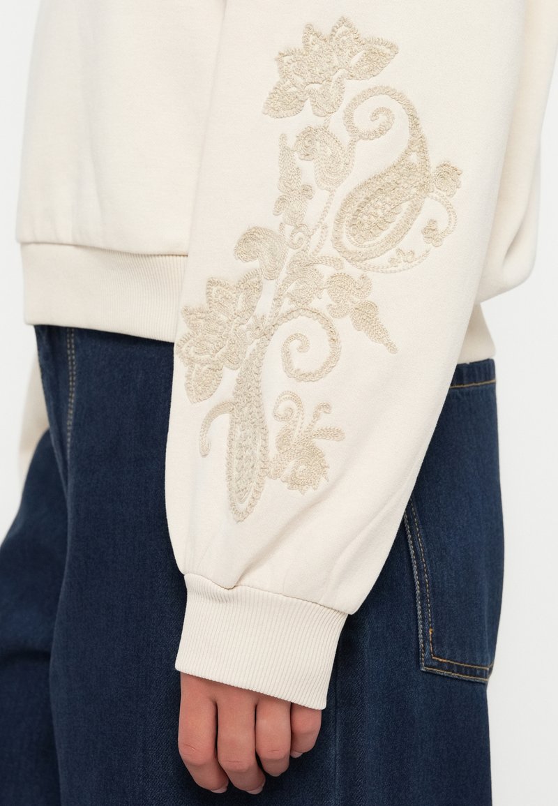 Beżowa bluza z ściągaczami, ozdobiona misternym beżowym haftem z motywami kwiatowymi na prawym rękawie. Połączona z ciemnymi dżinsami.