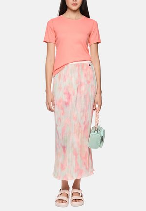 Frau trägt korallenfarbenes kurzes Oberteil, plissierten Maxi-Rock in Pastellrosa und Weiß, blassrosa Sandalen und hält eine kleine mintgrüne Handtasche.