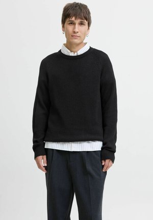 Pull en maille côtelée noir avec un col rond, des manches longues et une coupe oversize, superposé sur une chemise à col rayée blanche.
