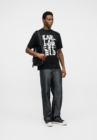 Νέος άνδρας με μαύρο γραφικό μπλουζάκι, σκούρο τζιν, μαύρα αθλητικά παπούτσια και μαύρη τσάντα ώμου στέκεται μπροστά σε απλό φόντο.