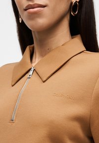 Γυναίκα που φοράει καφέ τοπ με φερμουάρ και γιακά με λογότυπο Calvin Klein, συνδυασμένο με χρυσούς κρίκους σκουλαρίκια.