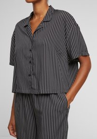 Camicia a maniche corte, a righe bianche e nere, con vestibilità rilassata e colletto piatto. Presenta una tasca e una texture liscia.
