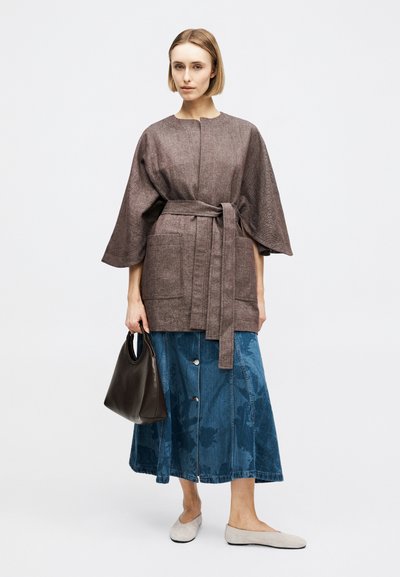 Γυναίκα που φοράει καφέ σακάκι με ζώνη και φαρδιά μανίκια, μπλε τζιν φούστα με κουμπιά, μπεζ φλατ παπούτσια, κρατώντας ένα σκούρο καφέ τσάντα.