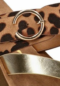 Sandalia con estampado de leopardo y una correa metálica dorada con detalle de hardware circular en oro. Parte superior de textura suave con un acabado brilloso contrastante.