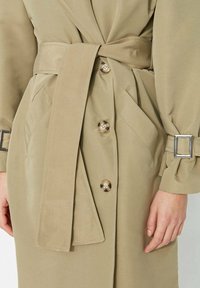 Jasno beżowy trencz z wiązanym paskiem w talii, zapinany na guziki, z ukośnymi bocznymi kieszeniami i mankietami zapinanymi na kwadratowe klamry.