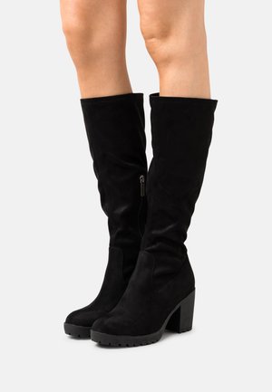 Bottes hautes en daim noir jusqu'aux genoux avec talons épais et fermetures éclair latérales, portées sur des jambes nues sur un fond clair uni.