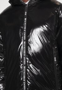 Schwarze glänzende Kapuzenjacke mit mehrfach gedrucktem "Versace Jeans Couture" auf dem zentralen Reißverschluss und den Reißverschlusszügen der Taschen.