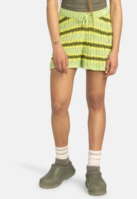 Stickade shorts i ljusgrön med gula och mörkgröna ränder, med elastisk midja och dragsnöre. Kombineras med gröna klompar och randiga strumpor.