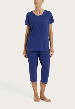 Donna con maglietta a maniche corte blu con tasca e pantaloni corti coordinati, in piedi a piedi nudi su uno sfondo semplice.