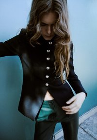 Črn po meri krojen suknjič s srebrnimi gumbi, z visokim ovratnikom in prilegajočo se silhueto. Nošen preko zelenih hlač z rahlim sijajem.