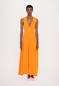 Orange maxi dress with a deep V-neck, sleeveless design, flowing silhouette, and a smooth texture; paired with pointed pale shoes.