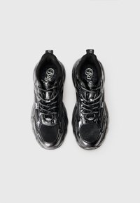 Sapatilhas atléticas pretas com acabamento brilhante, painéis de malha, atacadores e contornos de sola pronunciados. Identificação visível na lingueta e na lateral.