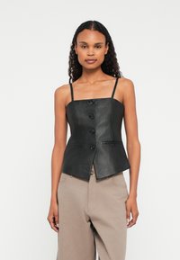 Naisel, kellel on pikad lokkis juuksed, on seljas must nööpidega nahast sansleeve top ja beežid püksid, seismas valge tausta ees.