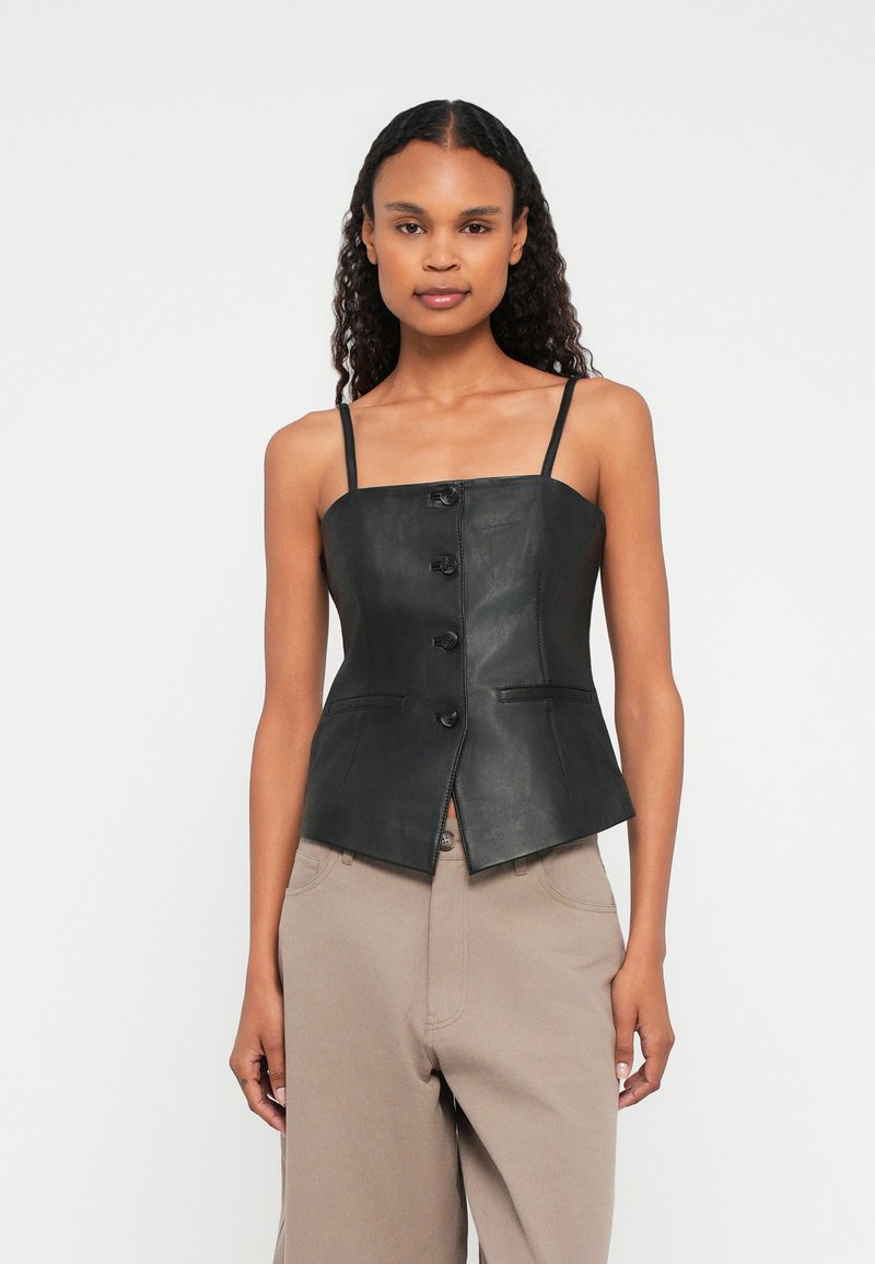 Naisel, kellel on pikad lokkis juuksed, on seljas must nööpidega nahast sansleeve top ja beežid püksid, seismas valge tausta ees.