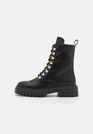 Botte de combat en cuir noir avec semelle épaisse, œillets dorés, lacets à l'avant et languette à l'arrière, vue de profil.