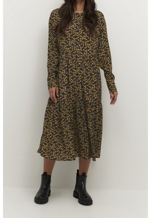 Femme portant une robe midi à manches longues, à motif floral sombre, et des bottines noires, debout devant un fond clair uni.