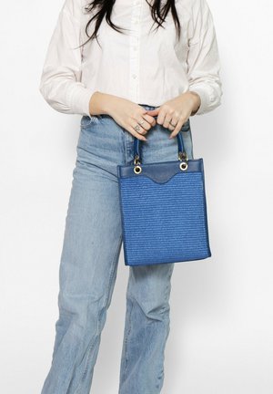 Vrouw in wit shirt en blauwe spijkerbroek die een gestructureerde blauwe handtas met dubbele handvatten vasthoudt voor een effen witte achtergrond.